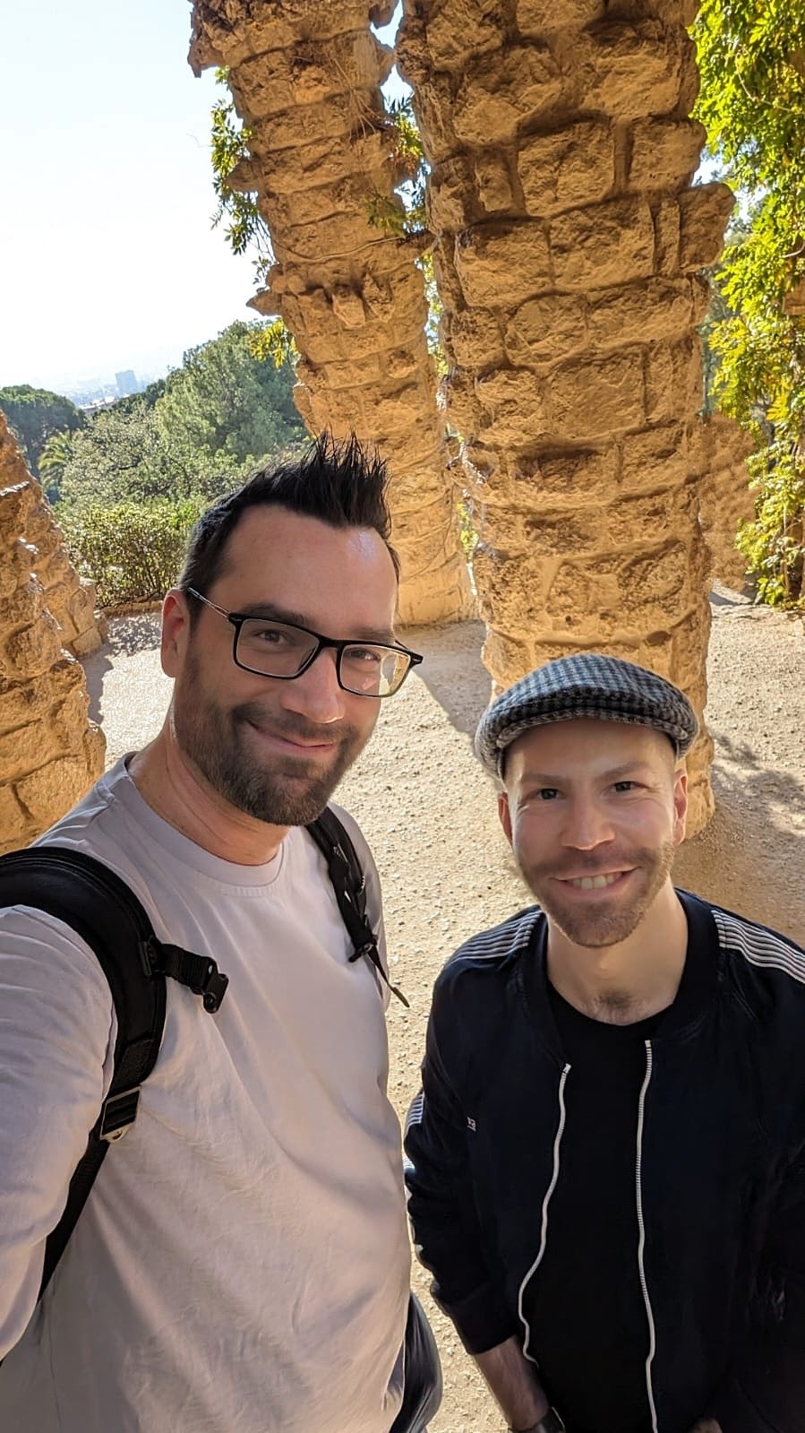 Podcast Hosts Toni und Sebastian lächeln auf einem Selfie. Im Hintergrund der Park Guell in Barcelona