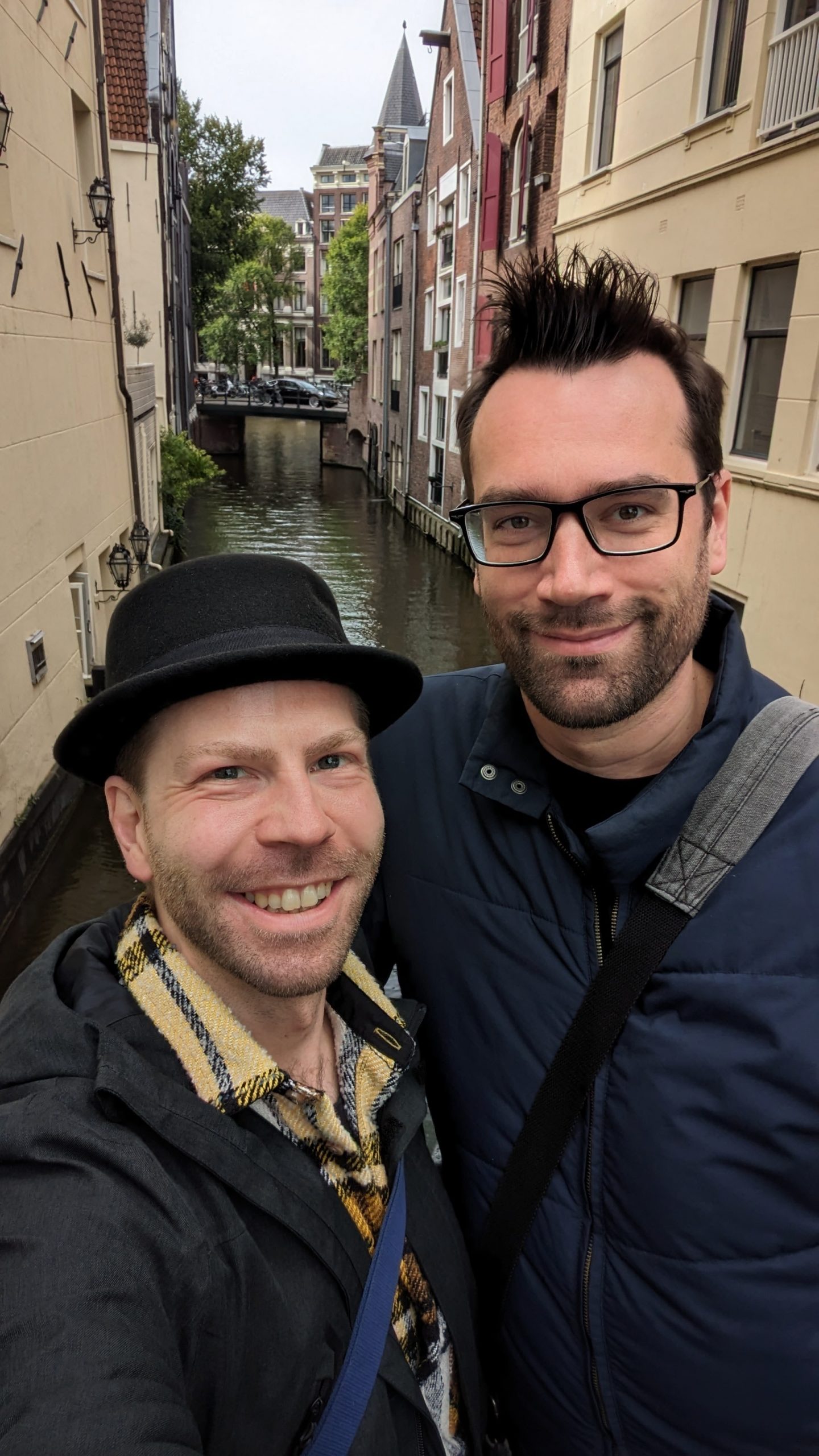 Toni und Sebi von Ausgang Podcast lächeln in die Kamera. Sie stehen vor einem der vielen Seitenarme eines Kanals in Amsterdam, die Sonne scheint.