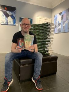 Fantasy Autor Christian Handel sitzt vor einem Stapel seines Buches "Rowan & Ash" und lächelt in die Kamera