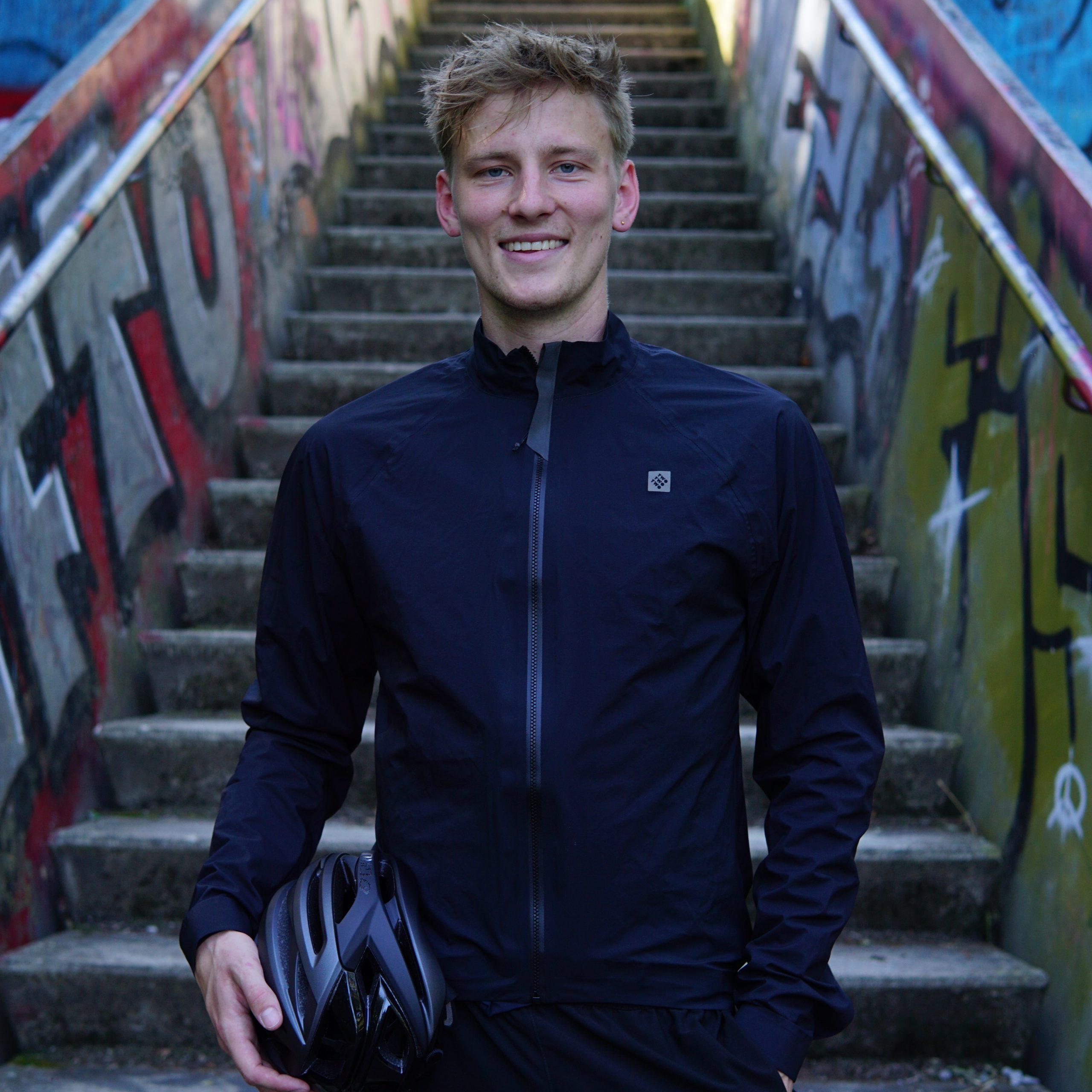 Fabian Abel aus Köln steht vor einer Treppe und lächelt in die Kamera. In der Hand hält er einen Fahrradhelm. Bei Ausgang Podcast erzählt von seiner Radtour die ihn über 25.000 Kilometer von Afrika nach Norwegen führen soll.