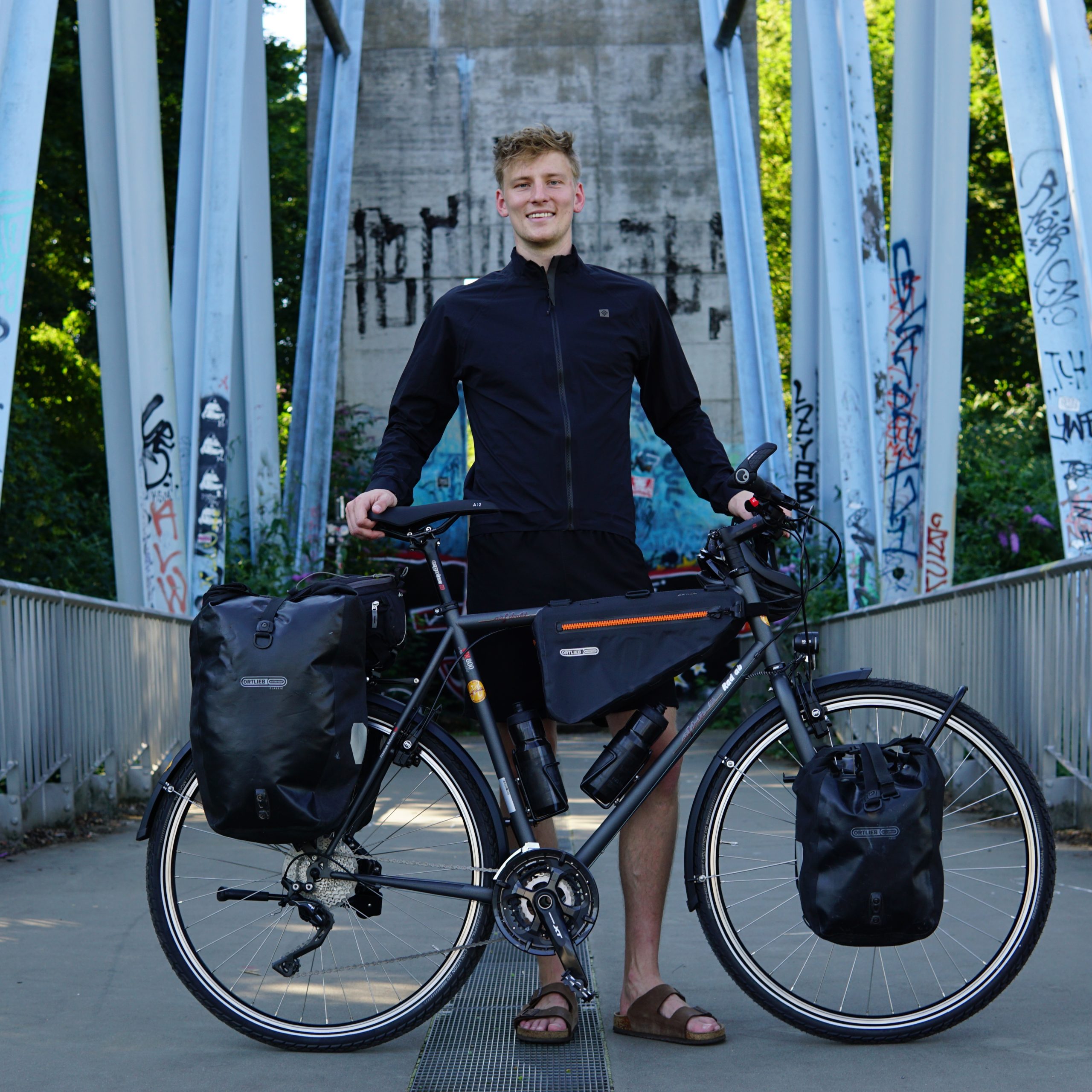 Fabian Abel aus Köln steht auf einer Brücke. Vor ihm steht sein Fahrrad mit dem er 25000 Kilometer von Südafrika nach Norwegen fahren möchte. Er lächelt in die Kamera.