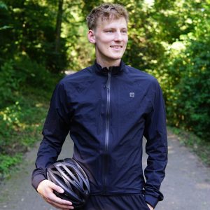 Fabian Abel aus Köln steht auf einem Weg im Wald. Im Hintergrund sind Bäume zu erkennen, auf die die Sonne scheint. Fabian schaut in die Ferne und hat seinen Fahrradhelm in der Hand. Er trägt eine blaue Fahrradjacke und lächelt leicht.