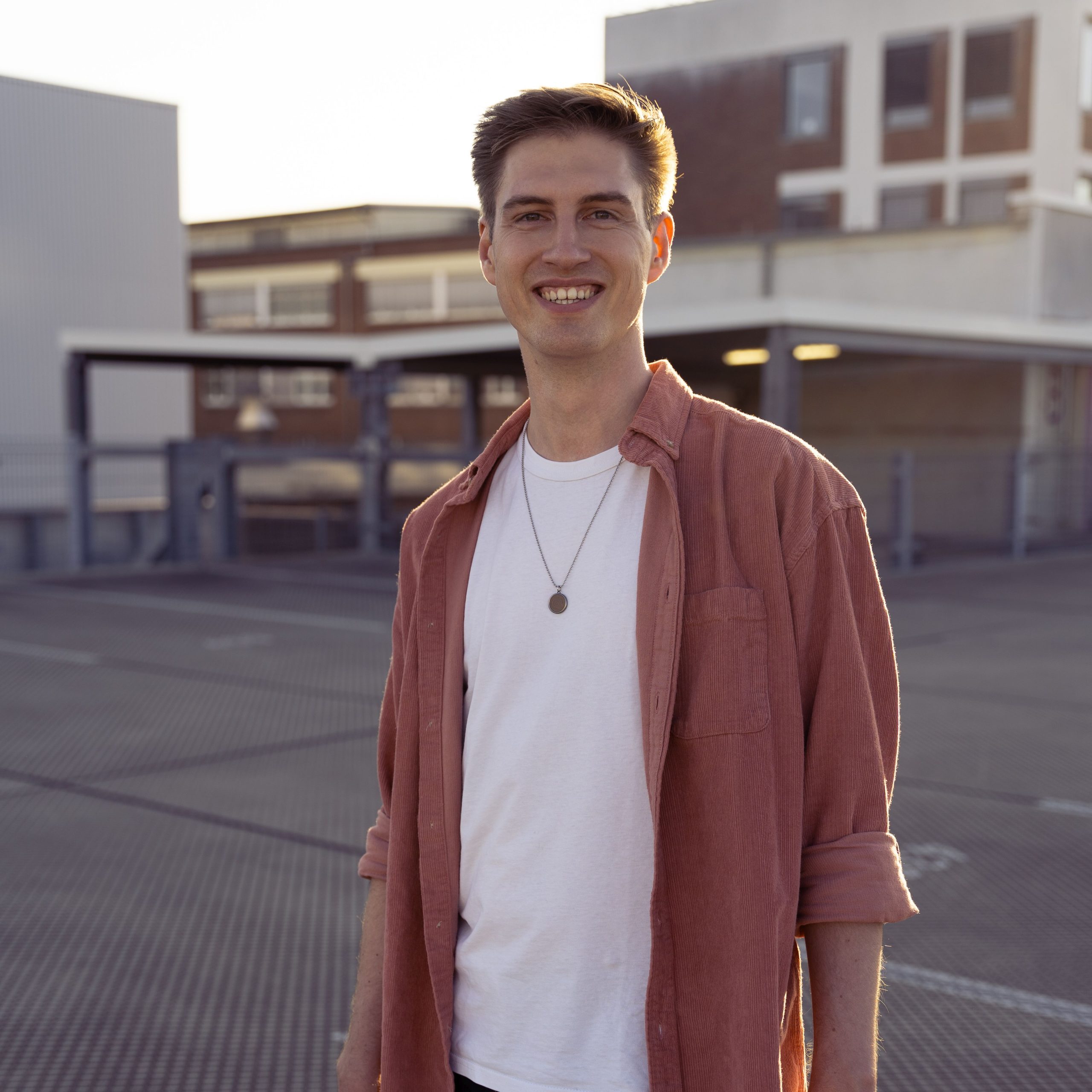 Radiologe Jan-Lucca Hennes aus Würzburg steht auf einer Betonfläche im Abendlicht. Die Sonne geht langsam unter und fällt seitlich auf ihn. Er lächelt in die Kamera und trägt ein rosa Hemd aus Cord und ein weißes T-shirt.