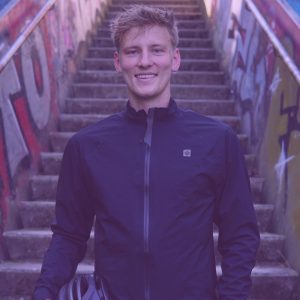 Fabian Abel steht mittig im Bild und lächelt direkt in die Kamera. Er steht vor einer steilen Treppe und hat einen Fahrradhelm in der Hand. Er trägt eine blaue Windjacke die bis zum Hals geschlossen ist.