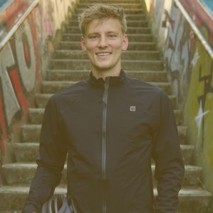 Fabian Abel steht mittig im Bild und lächelt direkt in die Kamera. Er steht vor einer steilen Treppe und hat einen Fahrradhelm in der Hand. Er trägt eine blaue Windjacke die bis zum Hals geschlossen ist.