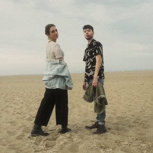 Frame the Moon sind ein Indie-Pop-Duo aus Köln. Es besteht aus Lena Schütten, links im Bild und Simon Schiller, rechts im Bild. Auf dem Foto stehen die beiden auf einem Strand. Lena hat braune Haare die zusammengebunden sind. Ihr Körper ist von der Kamera weggedreht, sie schaut aber genau in die Kamera. Sie tägt ein beigefarbenes Oberteil, eine schwarze Hose und schwarze Schuhe. Ihre Jeansjacke hat sie von den Schultern fallen lassen. Simon hat dunkelblonde kurze Haare und einen Bart. Er steht seitlich nach links zu Lena gedreht. Er trägt ein schwarzes Hemd mit kurzen Armen und beigem Muster. Er trägt eine dunkle Jeans die zum Knöchel hochgekrempelt ist und schwarze Schuhe. In der Hand hält Simon ebenfalls eine Jeansjacke. Das Foto hat der Fotograf Lukas Diekamp aufgenommen.