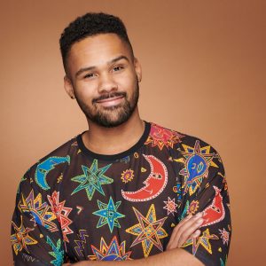 Das Foto zeigt den Diversity Marketing Coach Uchenna Jonas. Er hat die Arme leicht verschränkt und sieht mit verschmitzten Blick in die Kamera. Er hat kurze Haare und trägt ein dunkles Oberteil mit bunten Symbolen wie Mond und Sterne. Das Foto wurde aufgenommen von Marc Maria.