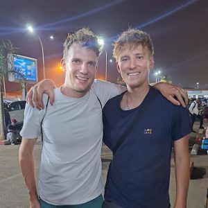 Fabian und Lucas Abel sind gemeinsam mit den Fahrrad durch Afrika gefahren. Das Foto zeigt Lucas Abel links im Bild und Fabian Abel rechts im Bild. Beide haben blonde Haare und tragen ein T-Shirt. Lucas T-Shirt hellgrau und Fabians T-Shirt ist dunkelblau. Die beiden stehen Arm in Arm und lächeln in die Kamera. Im Hintergrund ist zu erkennen, dass es dunkel ist. Das Bildrechte gehören Fabian Abel.
