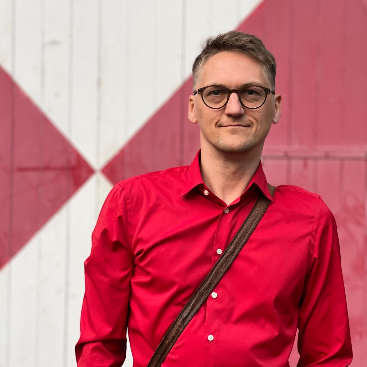 Das Bild zeigt eine männliche Person. Die Person hat blonde kurze Haare und trägt eine Brille. Sie lächelt in die Kamera. Die Person trägt ein rotes Hemd und eine braune Ledertasche schräg am Öberkörper. Das Foto zeigt den Kölner Künstler Maxim Probst.