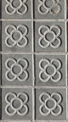 Das Bild zeigt einen Teil der vielen verzierten Bodenplatten der Gehwege in Barcelona. Diese sind berühmt für Muster und Ornamente. Auf diesem Bild ist ein Blumenmuster in jeweils einem von mehreren Quadraten zu erkennen.