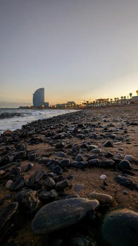 Das Bild zeigt das Hotel "W" in Barcelona im Sonnenuntergang. Es befindet sich im Hintergrund. Im Vordergrund ist der Strand mit einigen Steinen und einem Teil des Meeres zu sehen. Es sind Sonnenuntergang und alles liegt in der Abenddämmerung.